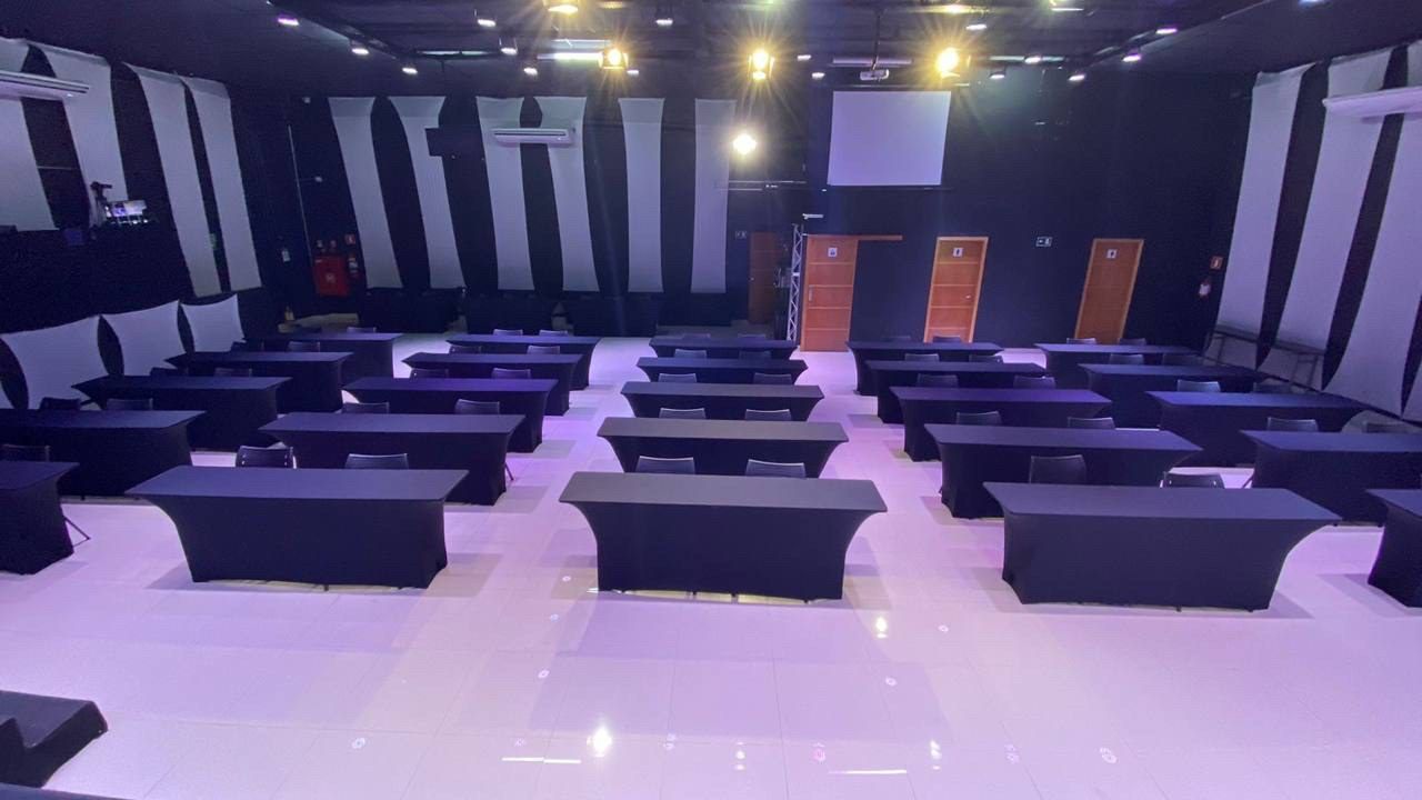 Espaço montado em formato escolar, com mesas de três lugares voltadas para o palco.
